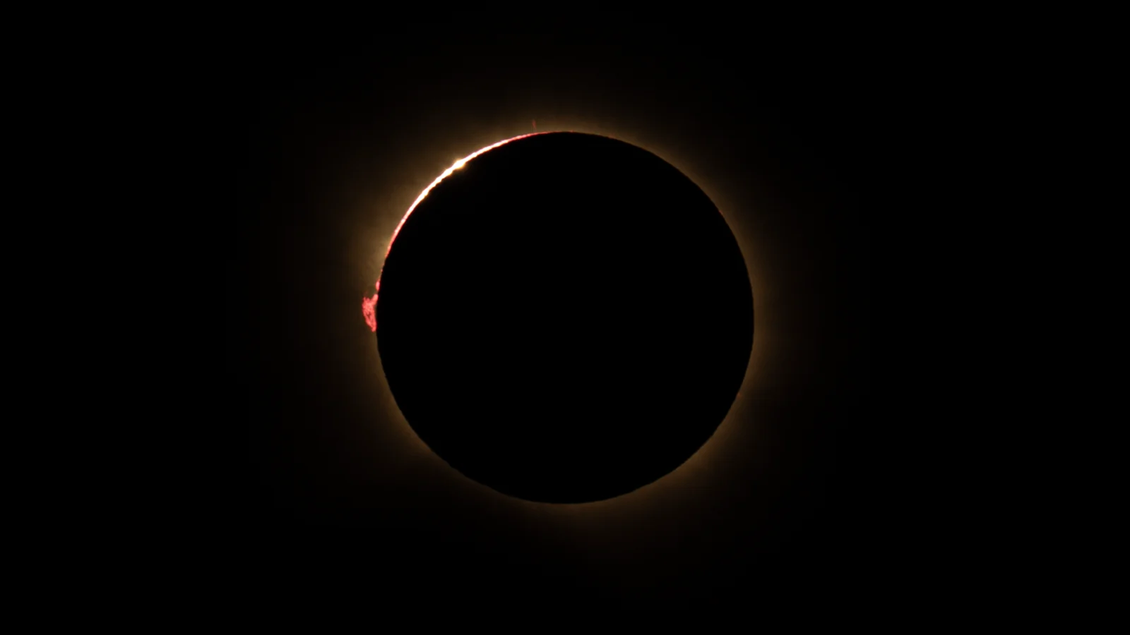 The total solar eclipse of 12 August 2026: the black disc of the Moon, a point of light at the upper left, a red prominence on the left edge.